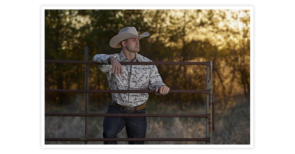 A man wearing Rodeo Felt Western hat in tan, posing at a gate looking away from teh camera.